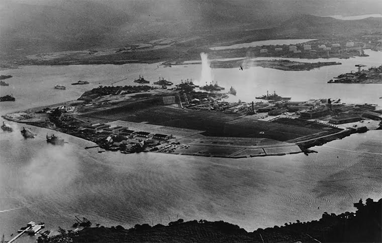 Photograph taken from a Japanese plane during the torpedo attack on ships moored on both sides of Ford Island. View looks about east, with the supply depot, submarine base and fuel tank farm in the right center distance. A torpedo has just hit USS West Virginia on the far side of Ford Island (center). Other battleships moored nearby are (from left): Nevada, Arizona, Tennessee (inboard of West Virginia), Oklahoma (torpedoed and listing) alongside Maryland, and California. On the near side of Ford Island, to the left, are light cruisers Detroit and Raleigh, target and training ship Utah and seaplane tender Tangier. Raleigh and Utah have been torpedoed, and Utah is listing sharply to port. Japanese planes are visible in the right center (over Ford Island) and over the Navy Yard at right. Japanese writing in the lower right states that the photograph was reproduced by authorization of the Navy Ministry. U.S. Naval History and Heritage Command Photograph.